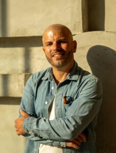 Cropped version - Love Coach Afterlove Olivier Laurent. Debout, souriant, soleil couchant. Habillé avec une chemise bleue, lunettes en poche