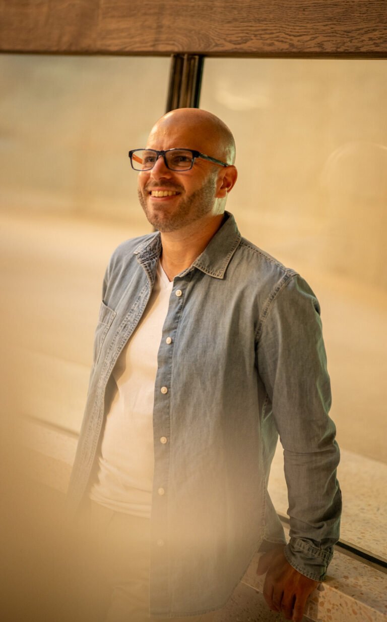 Love Coach Afterlove Olivier Laurent 3. Souriant, appuyé sur un rebord. Chemise bleue, t-shirt blanc. Décontracté